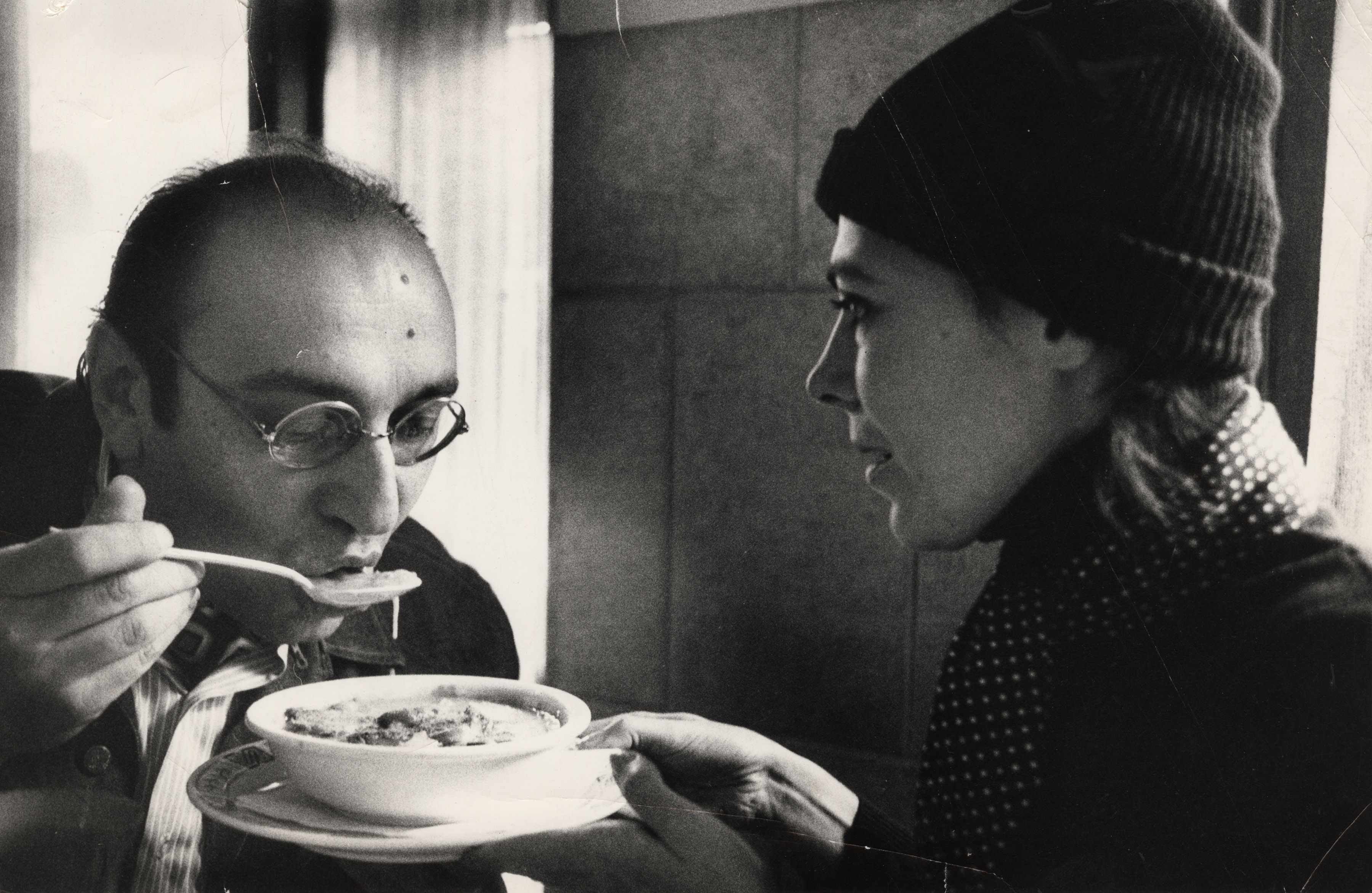 man with glasses eating soup while a woman in a hat holds the bowl
